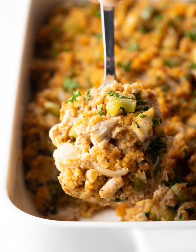 Metal spoon holding a serving of stuffing to camera. In the background is the white baking dish with crab dressing.