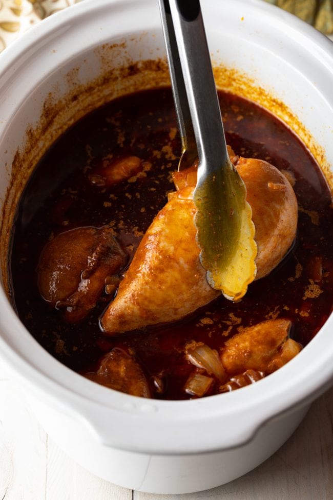 Tongs taking a chicken breast from the sauce in the crockpot to shred it. 