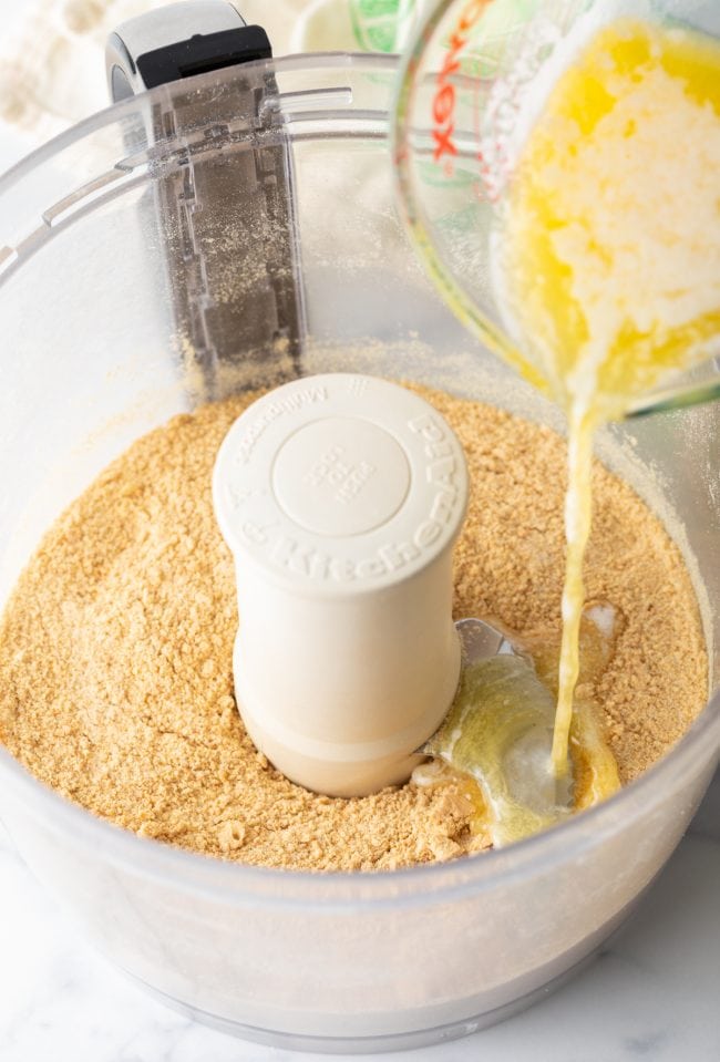 Graham cracker crumbs in a food processor with melted butter being poured on top.