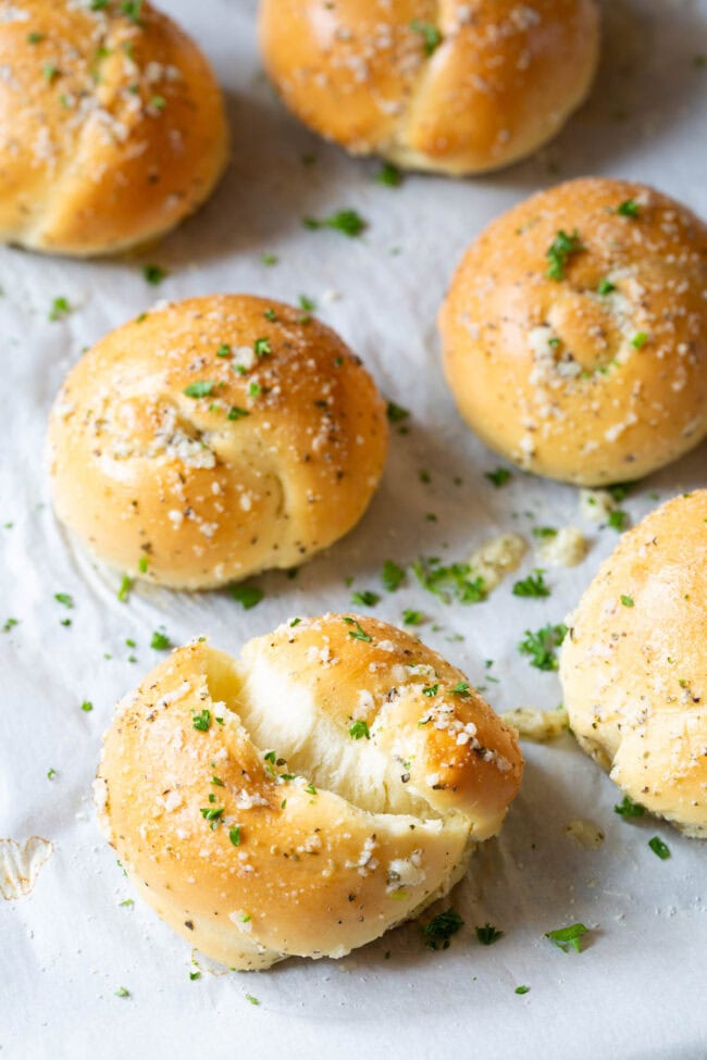 One garlic knot ripped to show inside with the rest behind it on a baking sheet. 