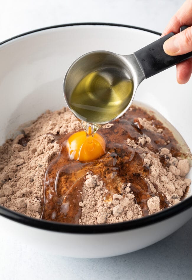 Hand with small measuring cup adding oil to the white measuring bowl with dry brownie mix and an egg.