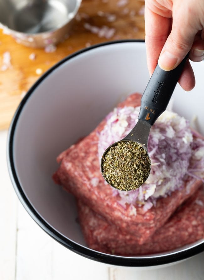 Ground lamb and onion in a mixing bowl with hand holding a measuring spoon of seasonings above it. 