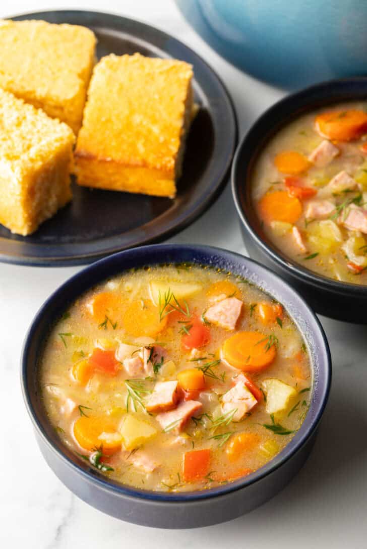 Two black bowls of ham and cabbage soup next to a black plate with 3 squares of cornbread.