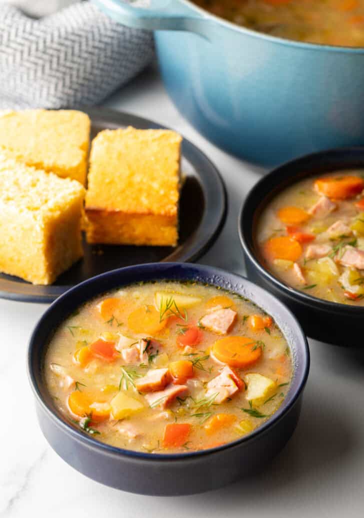 Two black bowls of ham and cabbage soup next to a black plate with 3 squares of cornbread.