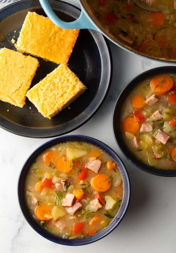 Top down view two black bowls of ham and cabbage soup next to a black plate with 3 squares of cornbread.