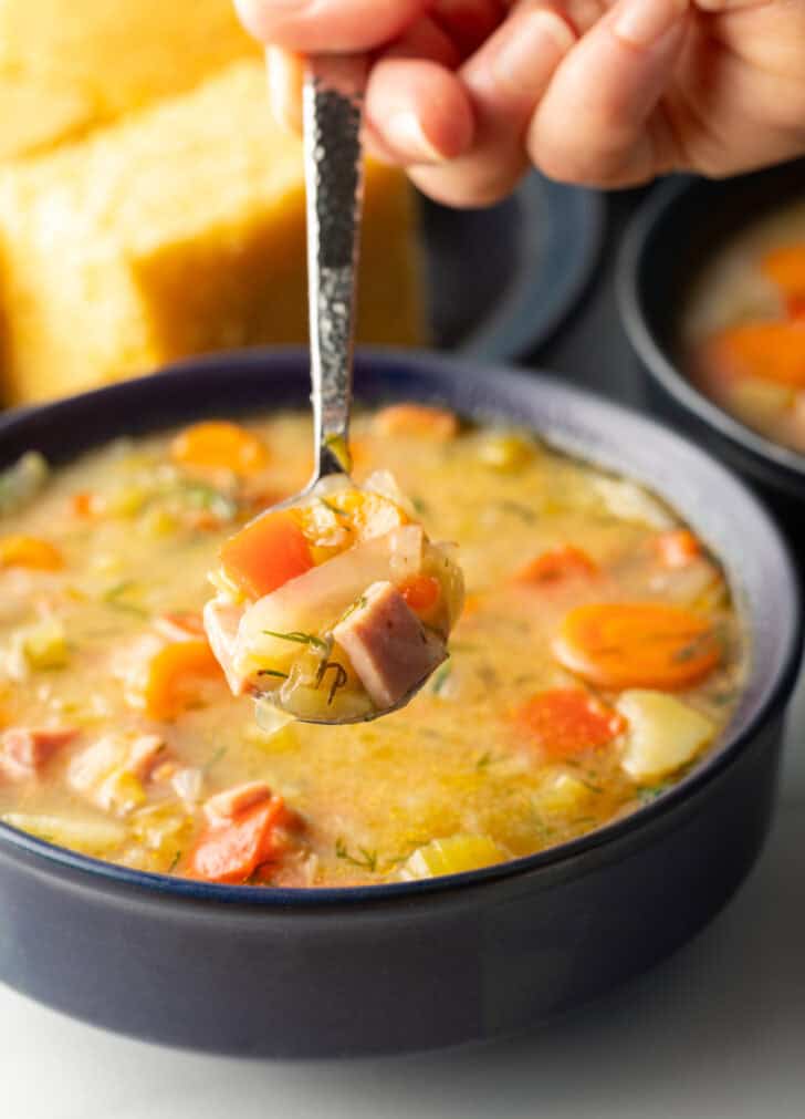 Spoonful of soup held over black bowl of cabbage and ham soup, showing to camera.