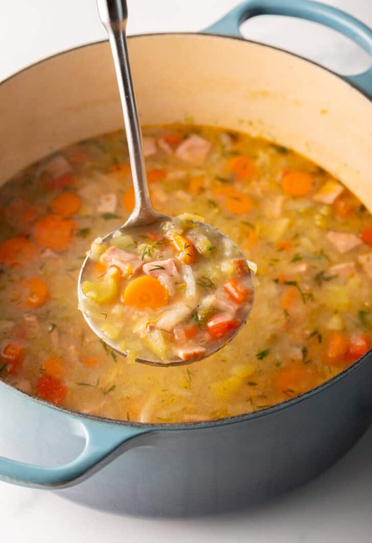 Metal ladle holding a scoopful of cabbage ham soup to camera, held over a blue cooking pot of soup.