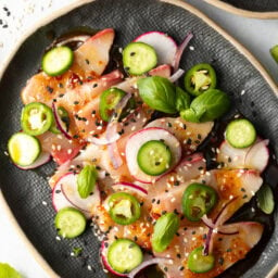 Top down view of black oval plate loaded with thin slices of yellowtail crudo topped with seasonings, thin slides of radishes, jalapenos, red onion, and a sprinkle of sesame seeds.