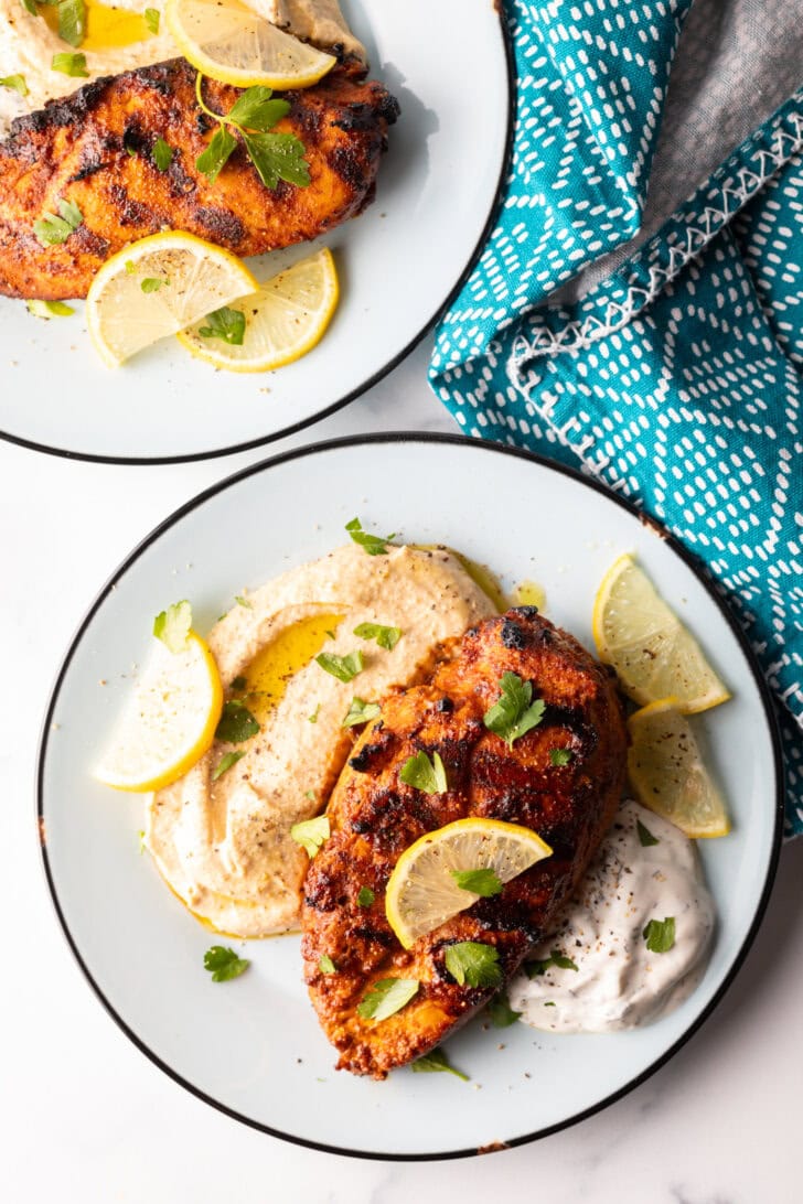 Top down Harissa baked chicken breast on a white plate, nestled between a serving of hummus and serving of Tzatziki sauce, and topped with slices of fresh lemon.