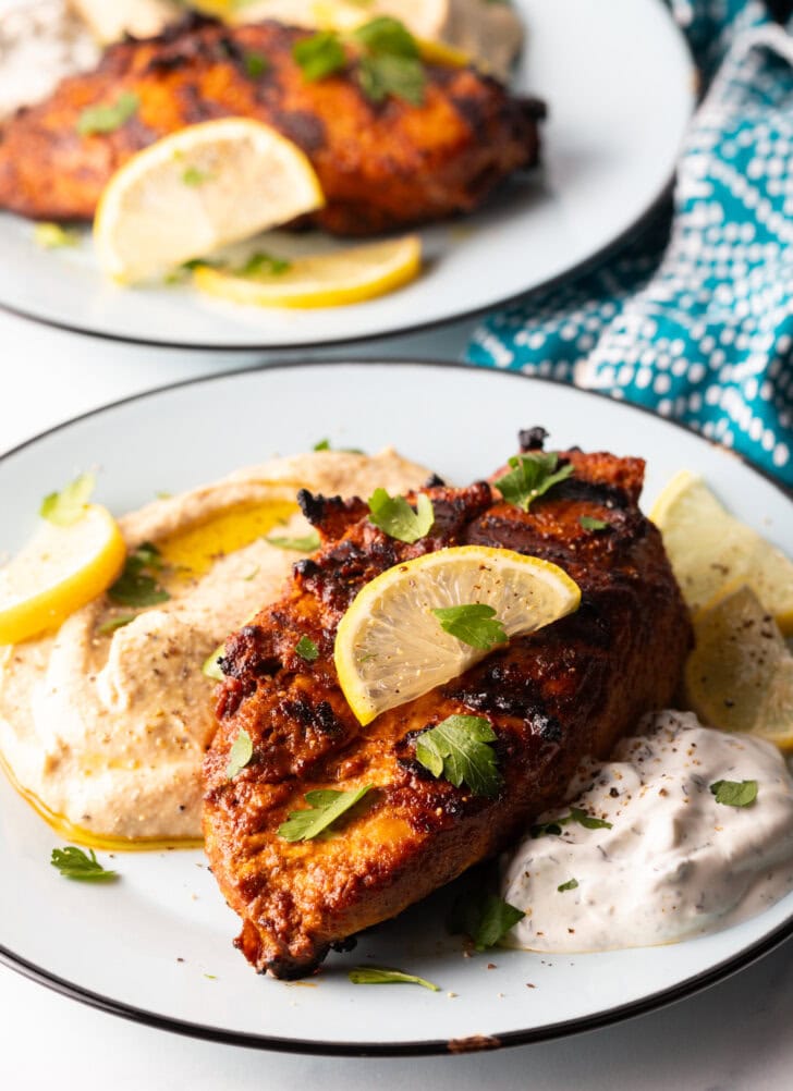 Harissa baked chicken breast on a white plate, nestled between a serving of hummus and serving of Tzatziki sauce, and topped with slices of fresh lemon.