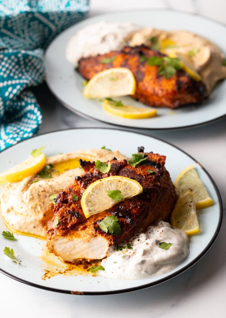 Harissa baked chicken breast on a white plate, nestled between a serving of hummus and serving of Tzatziki sauce, and topped with slices of fresh lemon. A bite has been removed from the chicken, showing the moist meat inside.