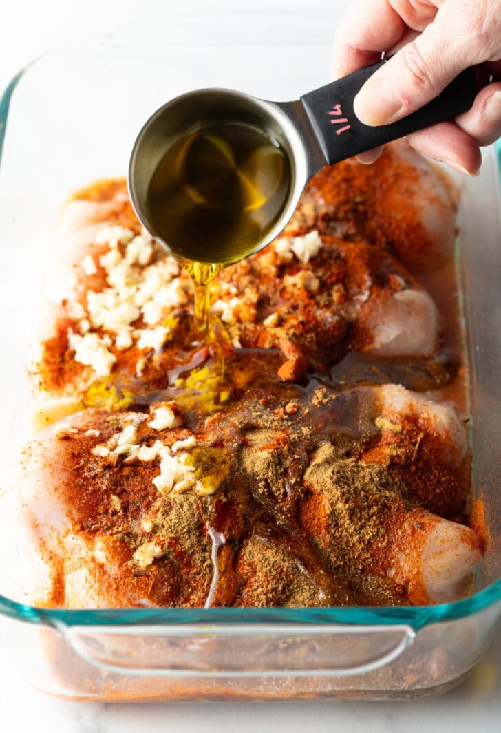 Hand adding oil from a measuring spoon to glass dish filled with raw chicken, spices, and chopped garlic.