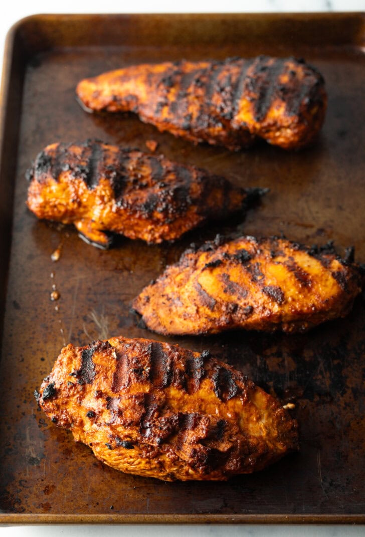 Four grilled chicken and harissa breasts on a metal baking sheet.