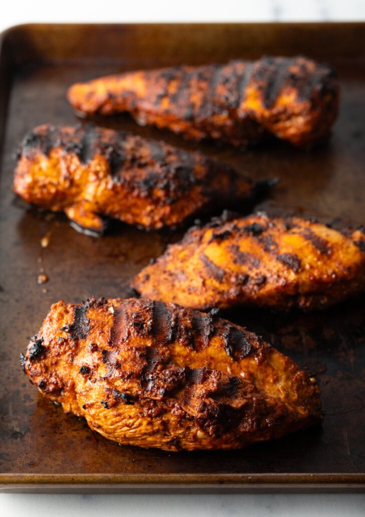 Four grilled chicken and harissa breasts on a metal baking sheet.