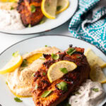 Harissa baked chicken breast on a white plate, nestled between a serving of hummus and serving of Tzatziki sauce, and topped with slices of fresh lemon.