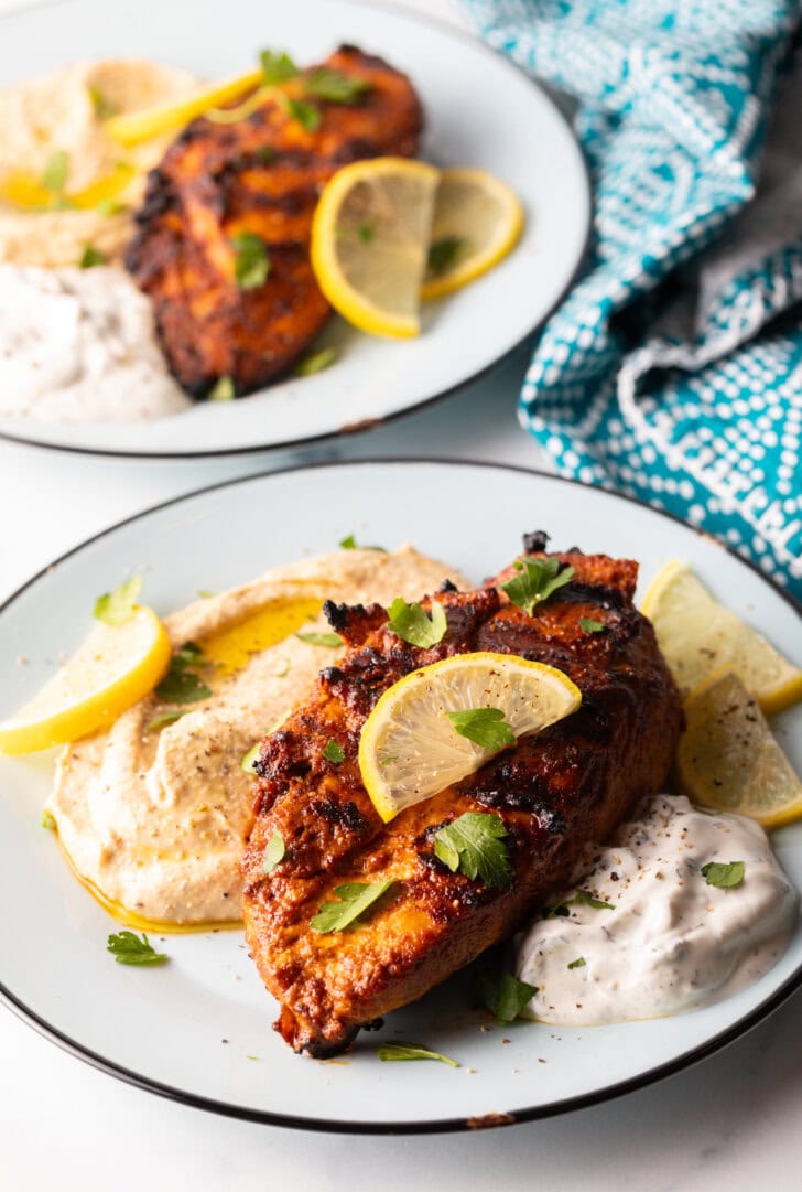 Harissa baked chicken breast on a white plate, nestled between a serving of hummus and serving of Tzatziki sauce, and topped with slices of fresh lemon.