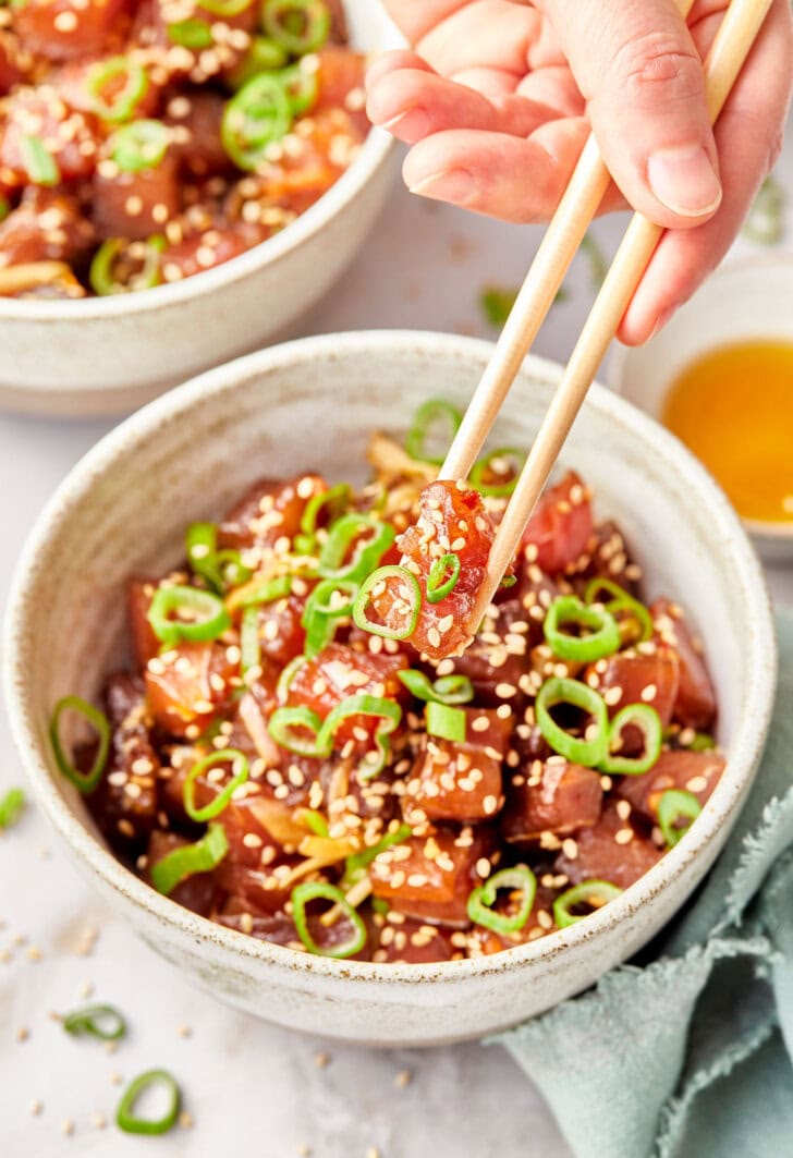 Hand holding a piece of raw tuna with chopsticks with more ahi poke in the background. 