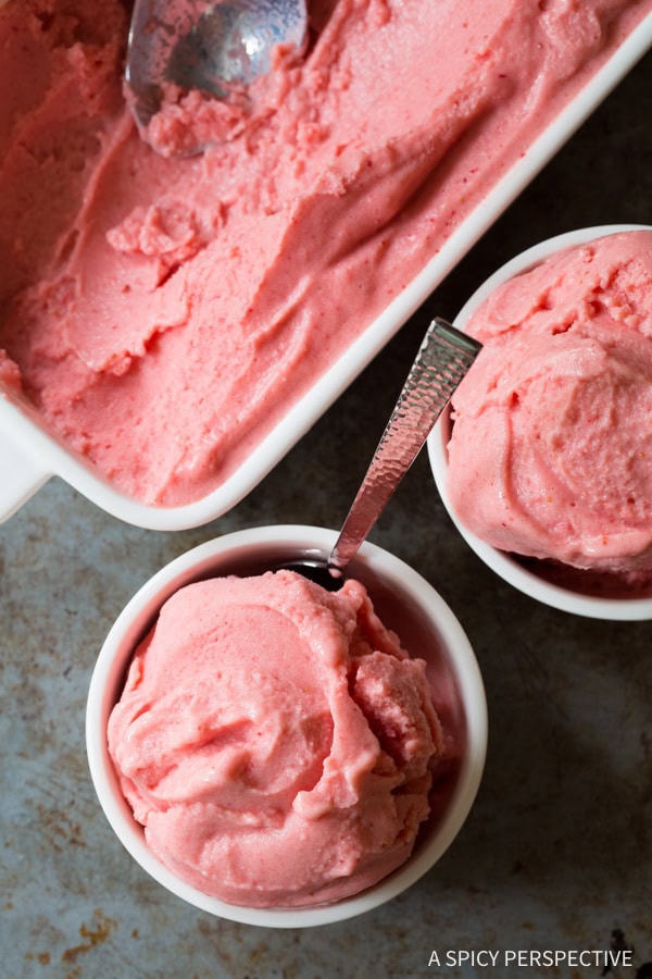 A top down view of a white bowl of strawberry sherbet with a metal spoon sticking out of it.