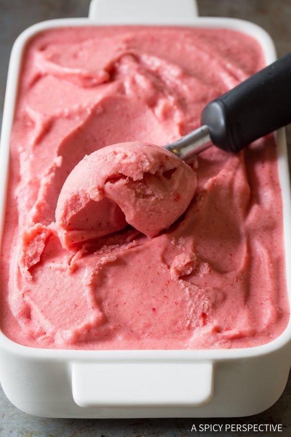 An ice cream scoop scooping strawberry pineapple sherbet out of a white serving dish.