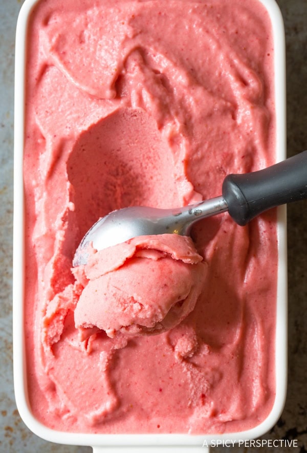 A top down view of an ice cream scoop scooping strawberry sherbet out of a white serving dish.