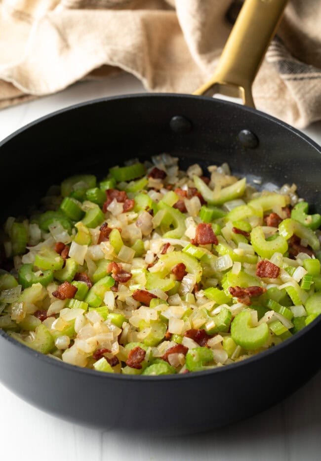 Sauteeing chopped celery, onions, and bacon in a black skillet.