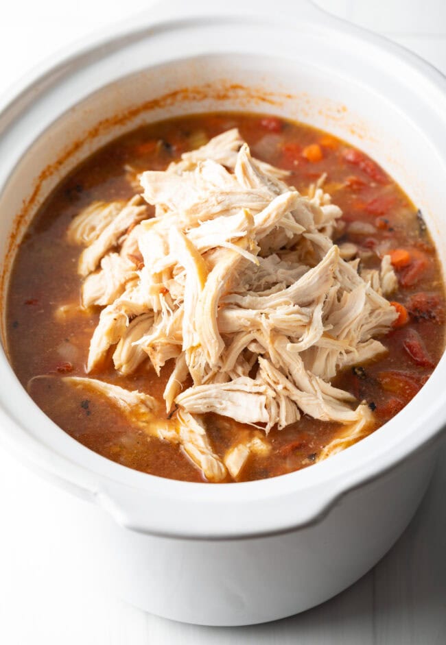 Shredded chicken pieces on top of tomato-based stew in white crock pot.