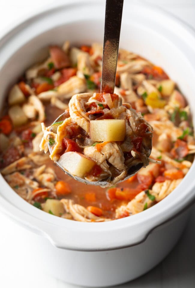 Metal ladle with stew showing to camera, white crock pot in the background.