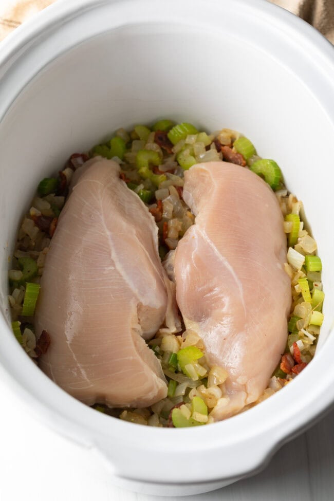 Top down view white slow cooker crock, with chopped sauteed veggies and two raw chicken breast on top.