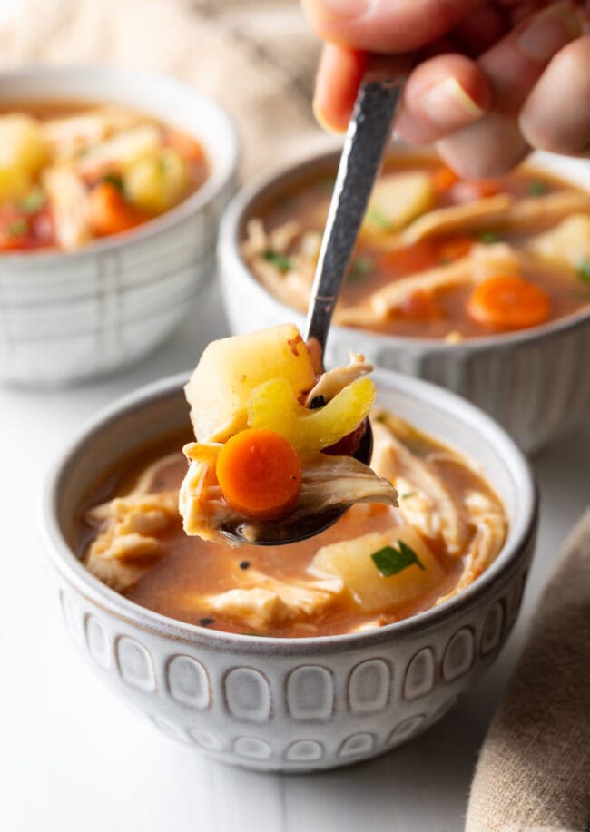 Hand with metal spoon, scooping serving of slow cooker chicken stew from one of three bowls.