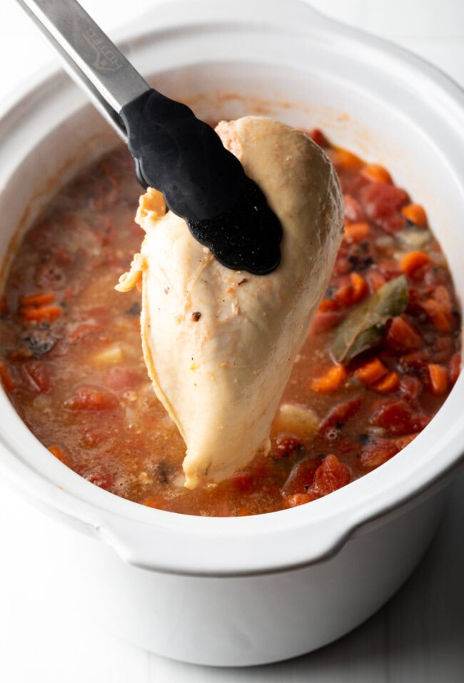 Tongs holding up cooked chicken breast to camera, above the crock pot with stew.