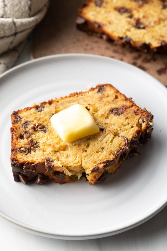 Protein Banana Loaf - Thick slice of healthy chocolate chip banana bread on a white plate, with a pat of butter on top.