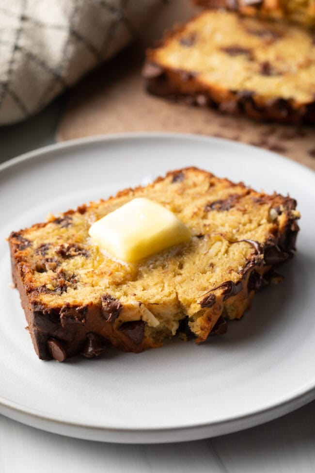 Thick slice of healthy chocolate chip banana bread on a white plate, with a pat of butter on top.