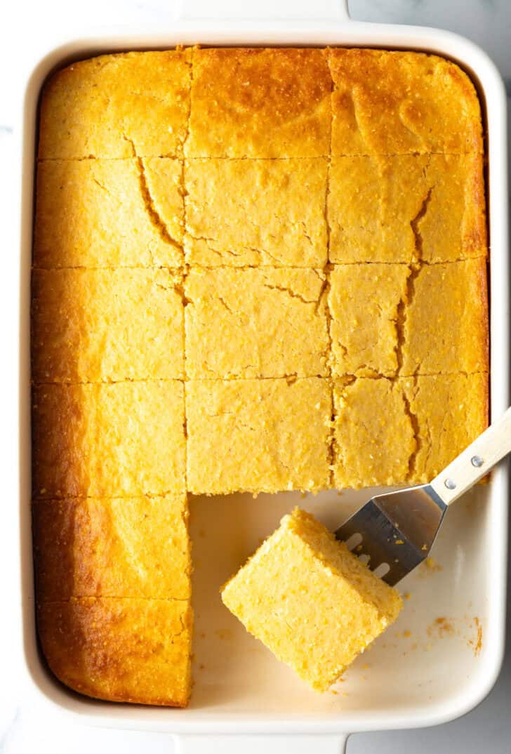 Top down view rectangle baking dish of homemade cornbread, cut into even squares. A metal spatula is removing a square of cornbread from the pan.