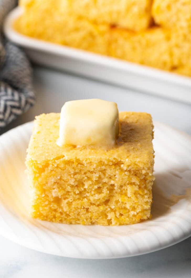 Square of fluffy sweet cornbread on a white plate, topped with a pat of butter.