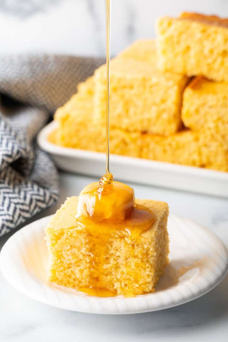 Square of cornbread is on a white plate, topped with a pat of butter being drizzled with honey.