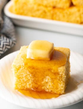 Square of cornbread is on a white plate, topped with a pat of butter and drizzled with honey.