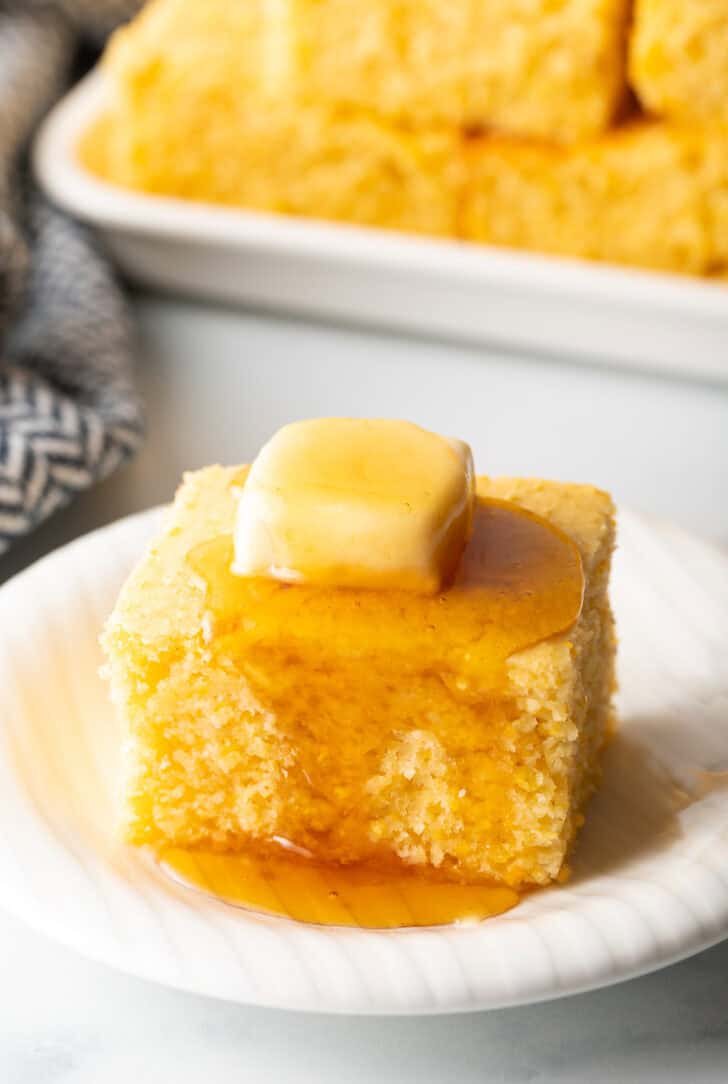 Square of sweet cornbread is on a white plate, topped with a pat of butter and drizzled with honey.