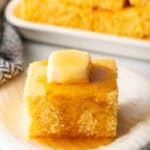 Square of cornbread is on a white plate, topped with a pat of butter and drizzled with honey.