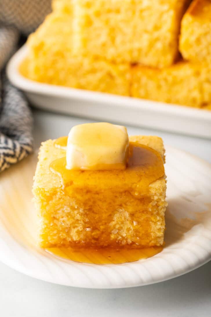 Square of cornbread is on a white plate, topped with a pat of butter and drizzled with honey.