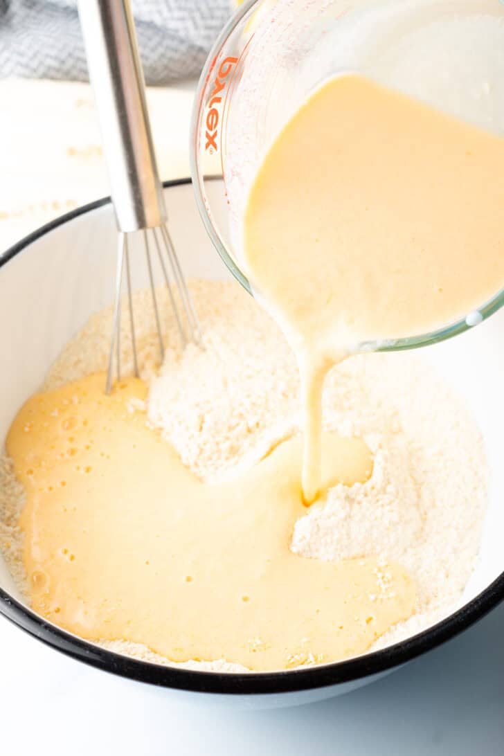 Adding milk from a glass measuring pitcher into a white mixing bowl with dry ingredients.