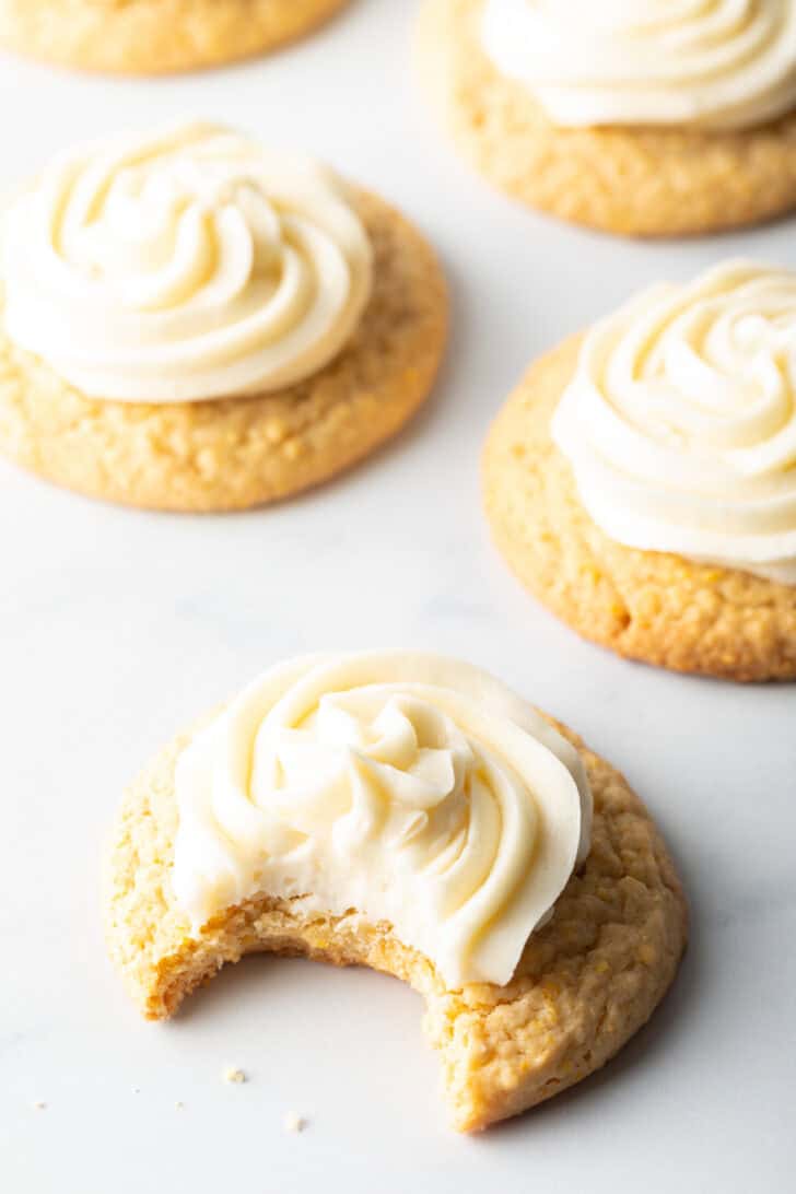 Several honey butter cornbread cookies on a white background, each topped with piped frosting. The cookie closest to camera has a bite taken out.