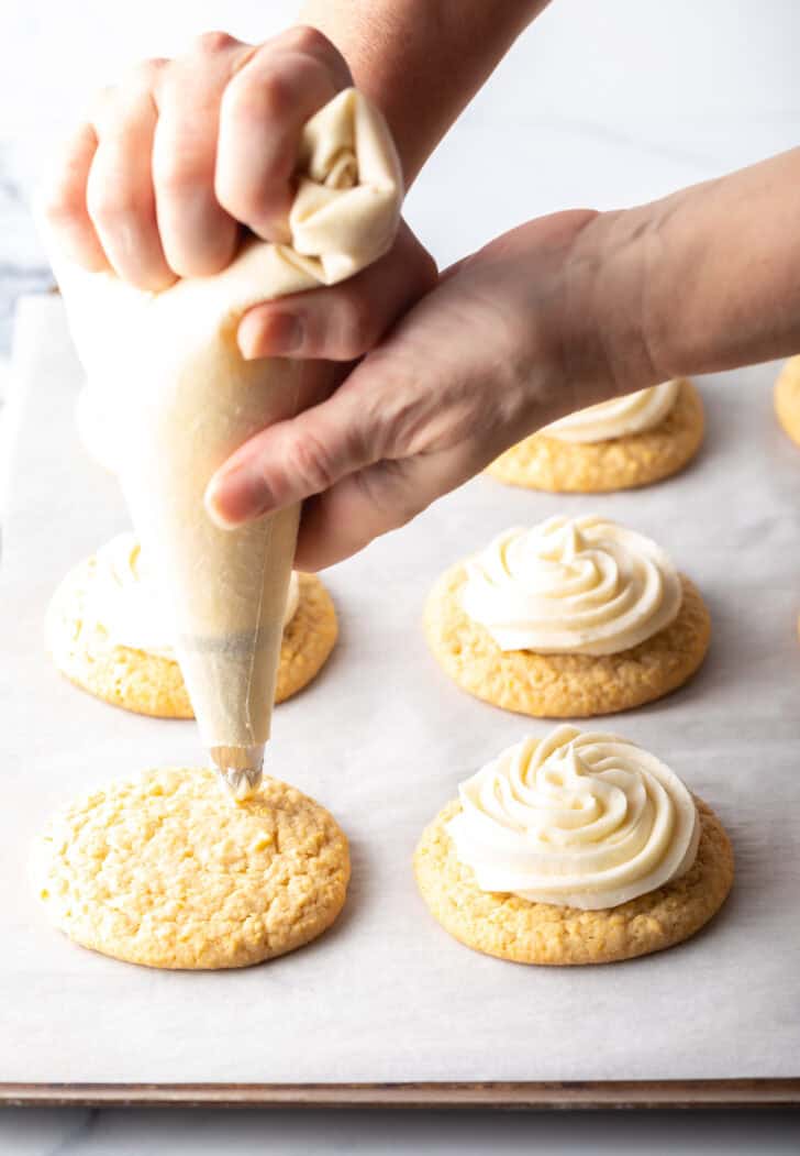 Hand with a piping bag piping white frosting onto the top of a cornbread cookie.