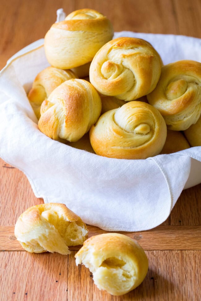 Dinner rolls in a linen lined bread basket with a dinner roll broken in half in front. 