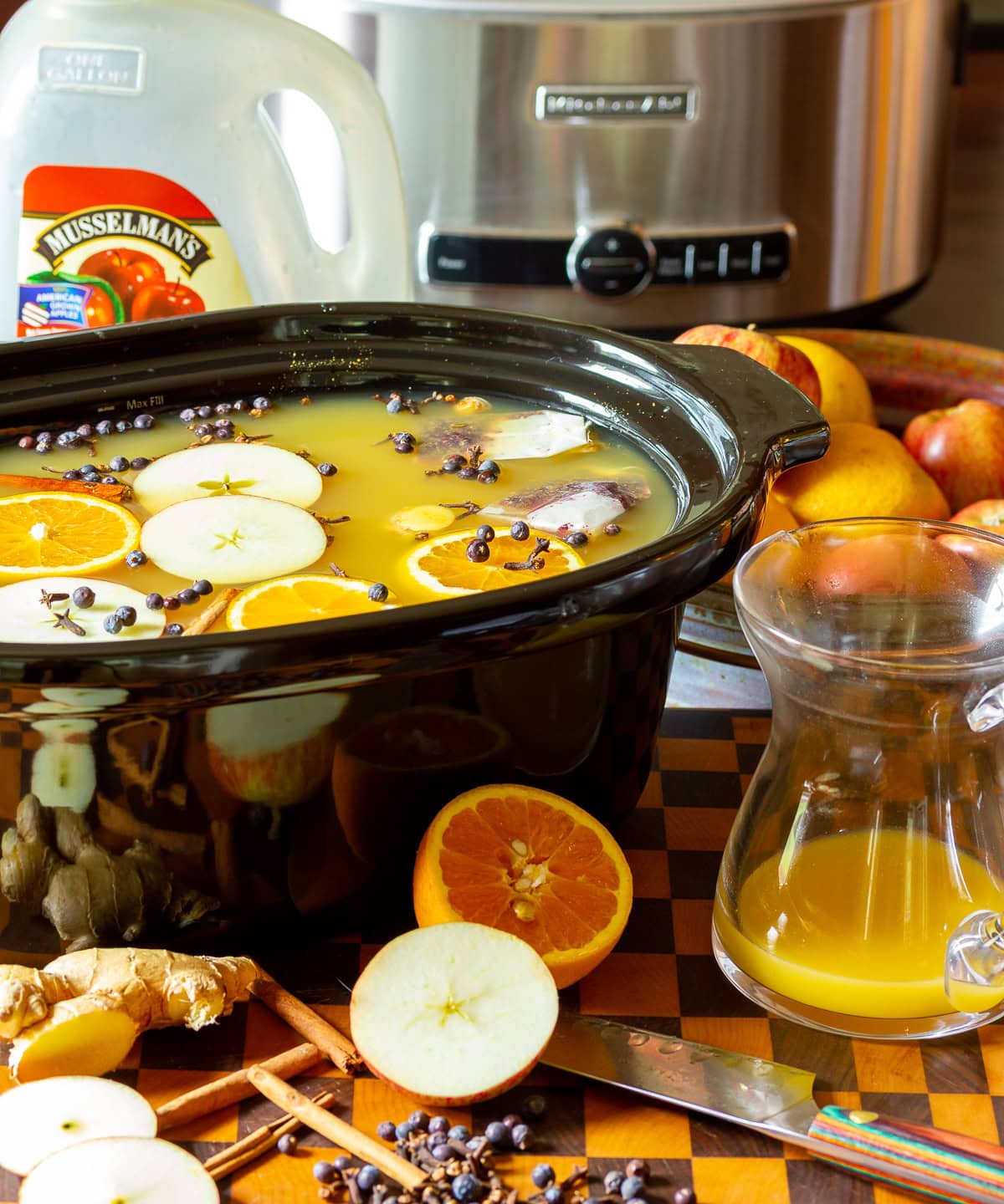 Apple cider wassail recipe in a crock pot with ingredients scattered in front. 