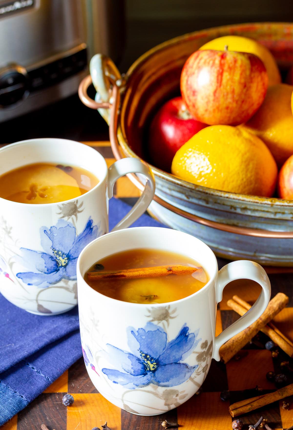 Two mugs of apple cider next to a bowl of oranges and apples. 