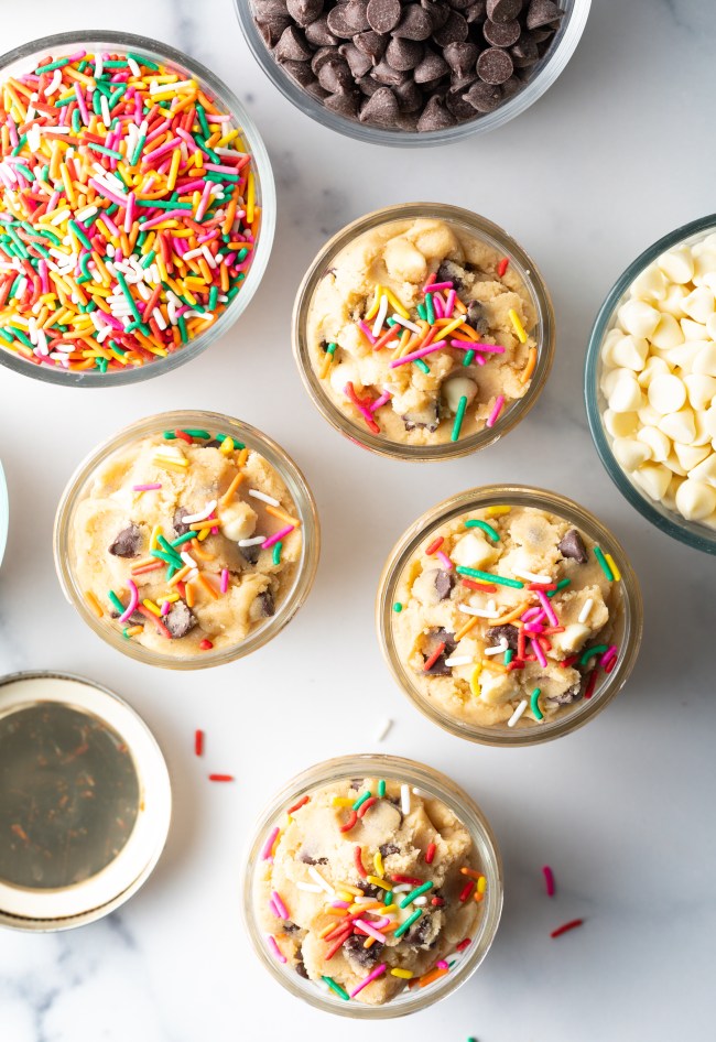 Top down four small glass containers with unbaked cookie dough topped with sprinkles. Also a glass bowl of sprinkles, and bowl of chopped nuts.