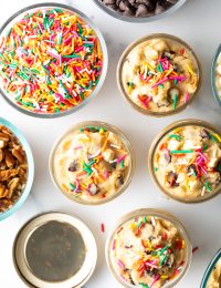 Top down four small glass containers with edible cookie dough topped with sprinkles. Also a glass bowl of sprinkles, and bowl of chopped nuts.