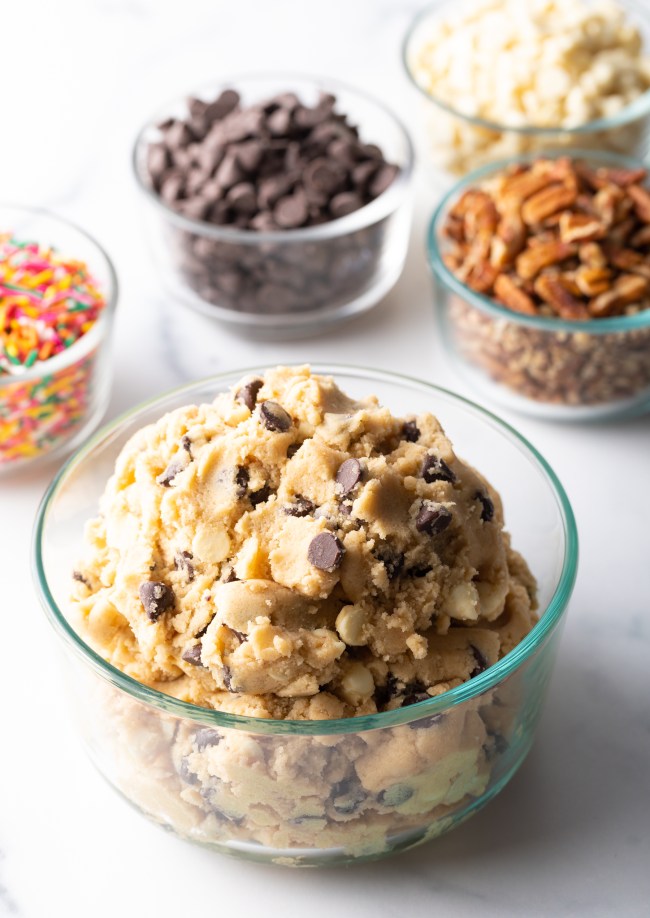 Glass container with chocolate chip cookie dough. In the background are small glass dishes with chocolate chips, sprinkles, and chopped nuts.
