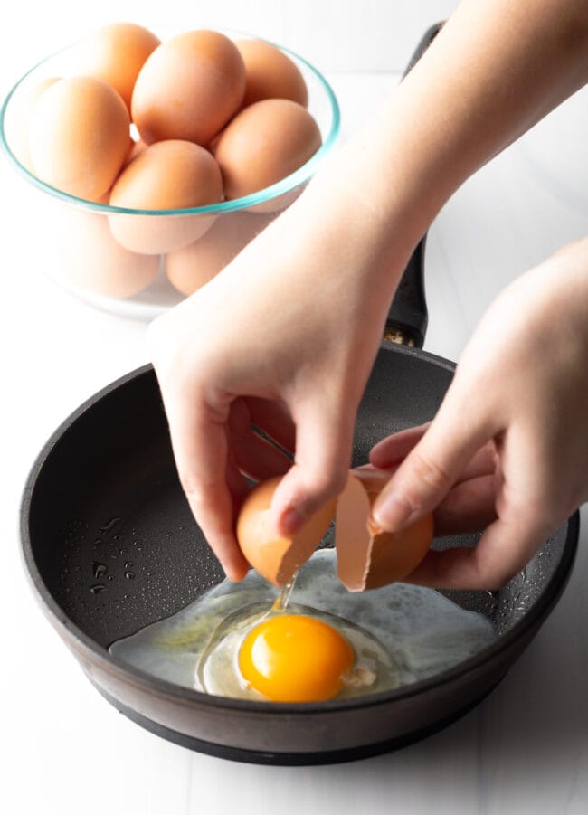 Hands cracking an egg just above a black frying pan.