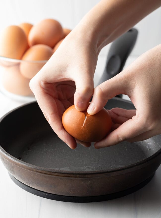 Hands cracking an egg just above a black frying pan.